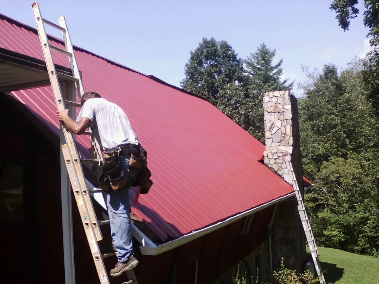 Roofer on a ladder beside a red metal roof and stone chimney — Soffit & Fascia Repair by Roofing Brothers in Aroda